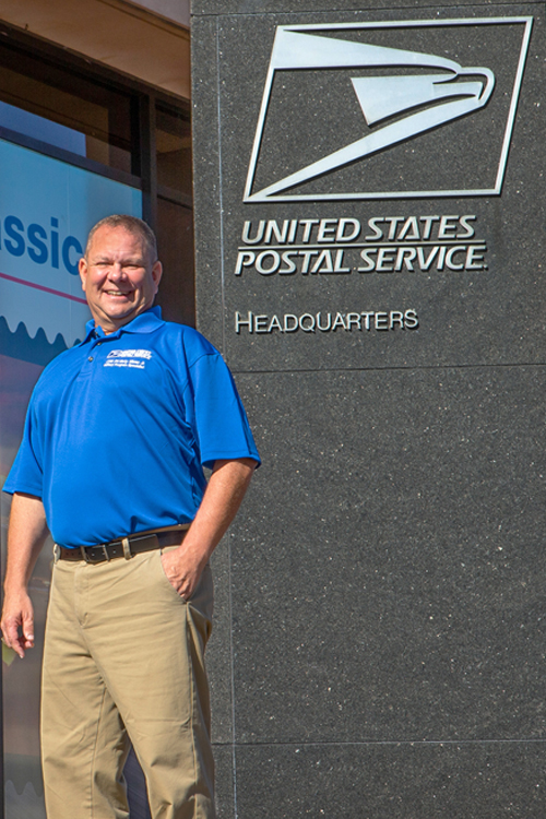 A man wearing a blue polo shirt with the USPS logo stands next to the USPS headquarters building sign.