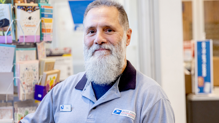 A man with gray hair and a white beard smiles while standing in a Post Office retail space. He wears a Postal Service uniform shirt and is seen from the chest up.