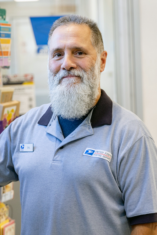 A man with gray hair and a white beard smiles while standing in a Post Office retail space. He wears a Postal Service uniform shirt and is seen from the chest up.