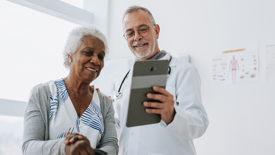 An older woman sits in a medical examining room, smiling as a doctor in a lab coat shows her a digital tablet