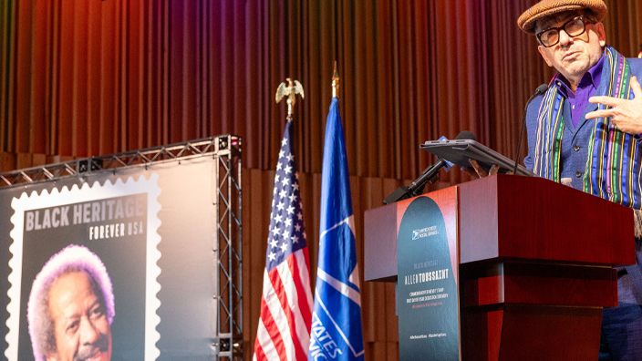 Elvis Costello, dressed in a blue suit, stands at a lectern and gestures near an oversized poster of the Allen Toussaint postage stamp