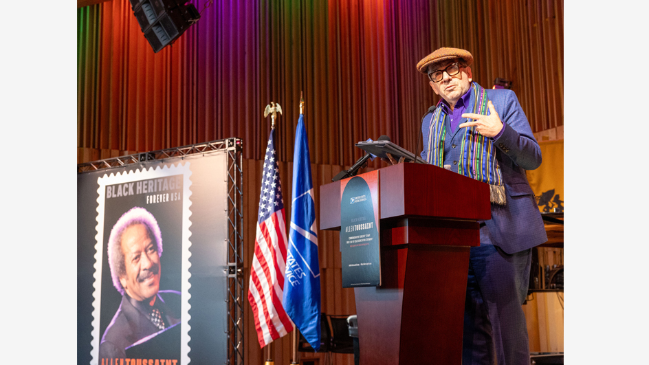 Elvis Costello, dressed in a blue suit, stands at a lectern and gestures near an oversized poster of the Allen Toussaint postage stamp