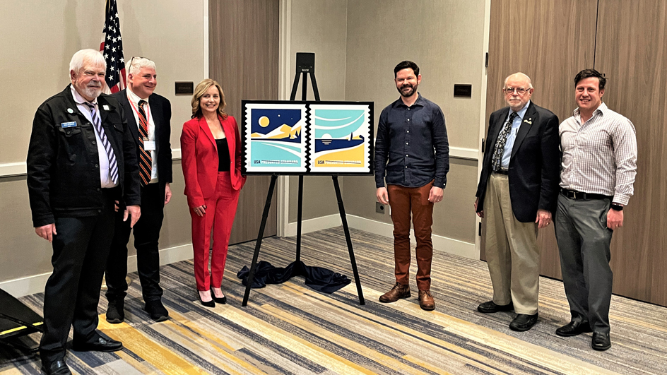 A group of five men and one woman stand next to an enlarged image of the American Vistas stamps.