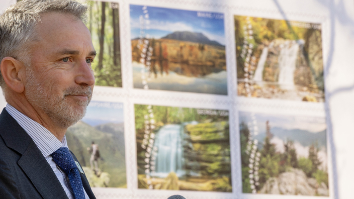 A man in a business suit stands at a lectern in a forested setting, near a large poster displaying stamp images