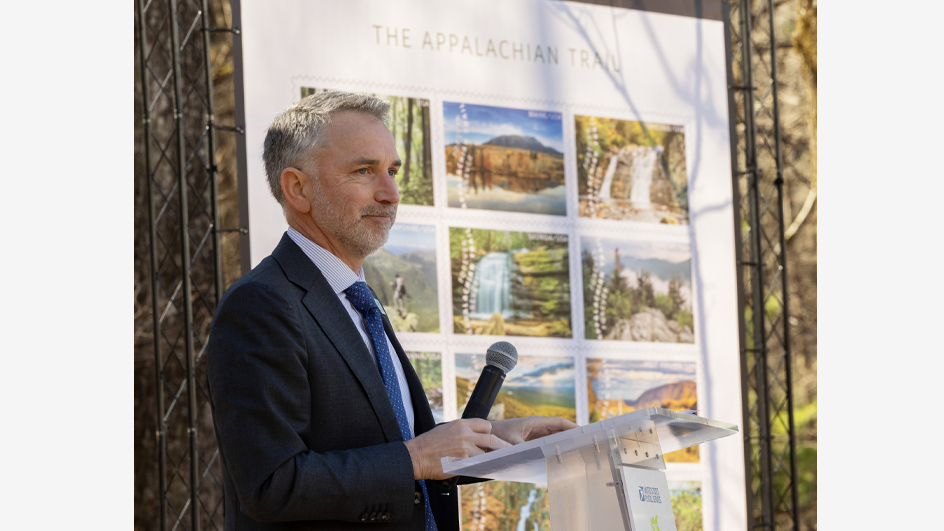 A man in a business suit stands at a lectern in a forested setting, near a large poster displaying stamp images