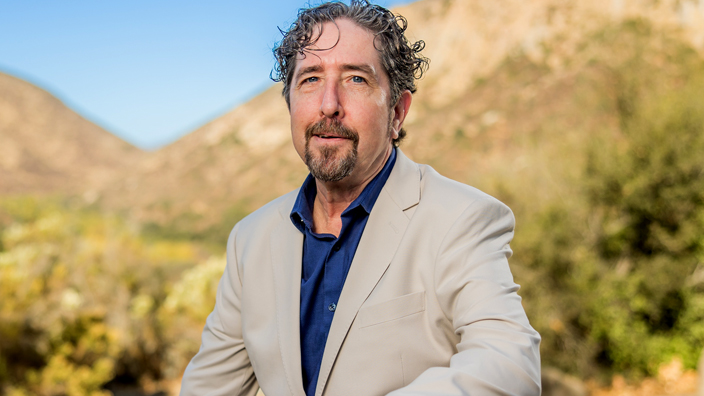 A man wearing a beige suit sits on a rock in a desert setting