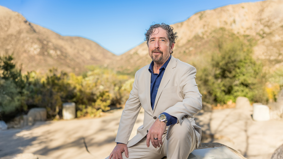 A man wearing a beige suit sits on a rock in a desert setting