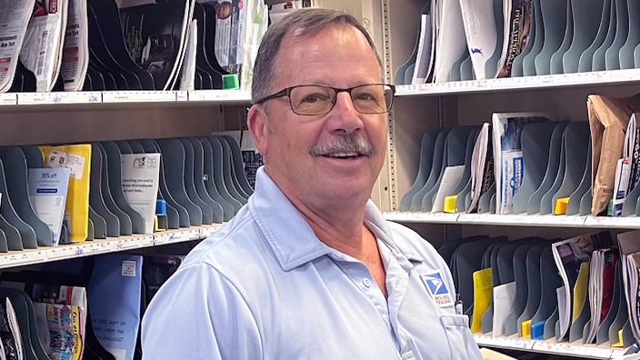 A man in a light blue USPS shirt smiles while standing in front of a sorting rack.