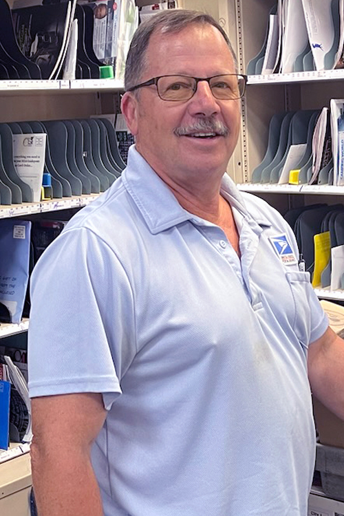 A man in a light blue USPS shirt smiles while standing in front of a sorting rack.