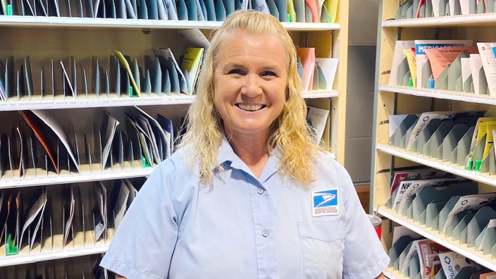 A woman wearing a Postal Service uniform shirt smiles brightly while standing in a Post Office workroom