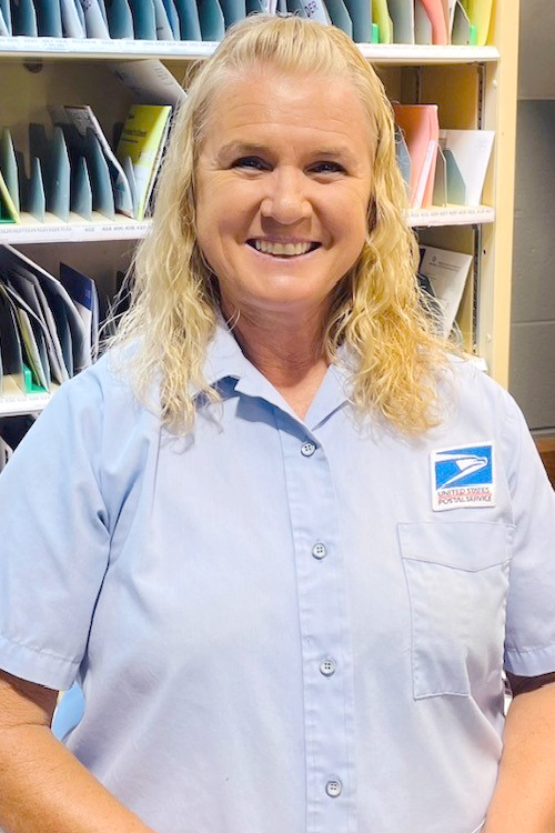 A woman wearing a Postal Service uniform shirt smiles brightly while standing in a Post Office workroom