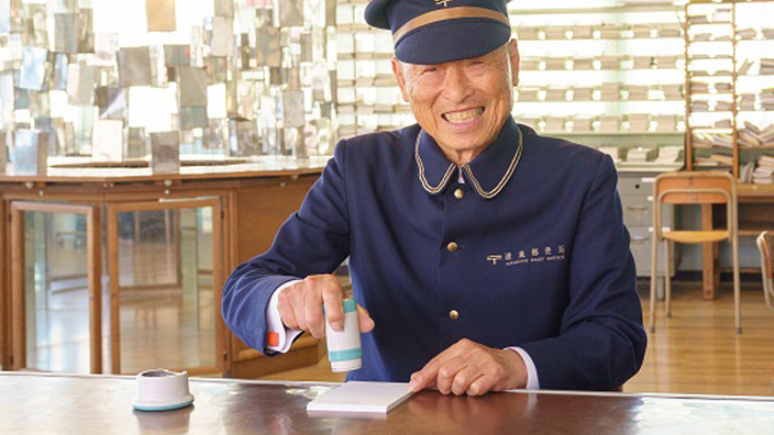 A Japanese man in a sharp postal uniform stands at a counter and smiles brightly