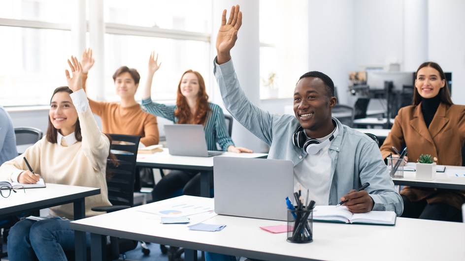 A group of young adults sit in a classroom and watch as a fellow student eagerly raises his hand to speak