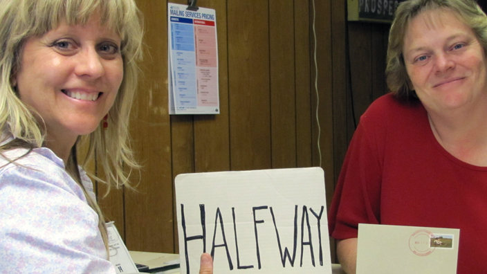 A smiling woman stands at a Post Office counter, and another woman stands on the other side