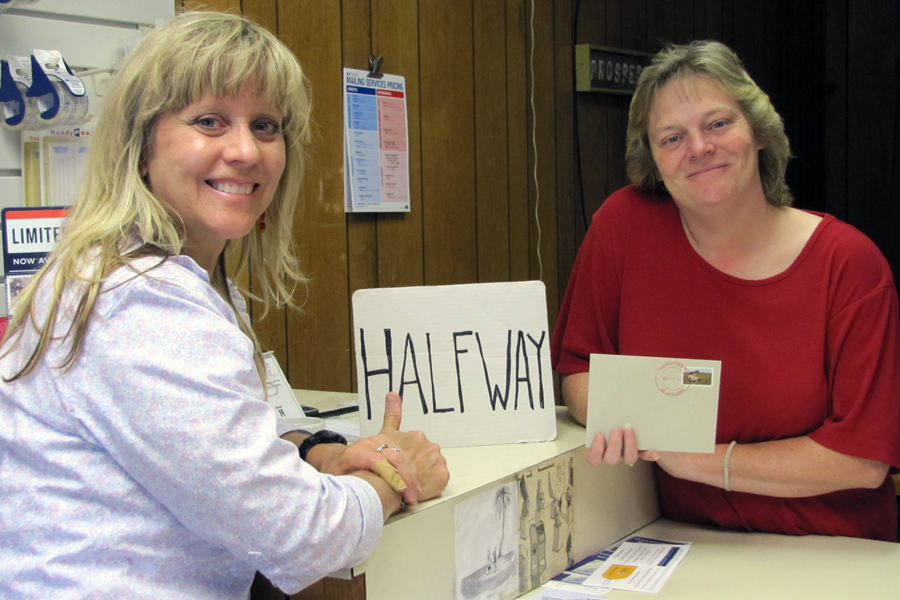 A smiling woman stands at a Post Office counter, and another woman stands on the other side