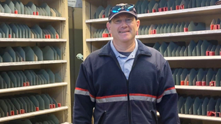 A smiling man wearing a Postal Service uniform stands in a Post Office workroom