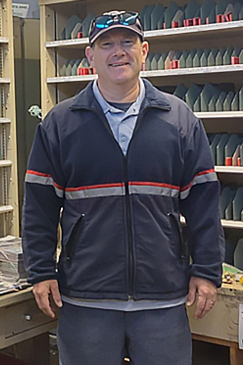 A smiling man wearing a Postal Service uniform stands in a Post Office workroom