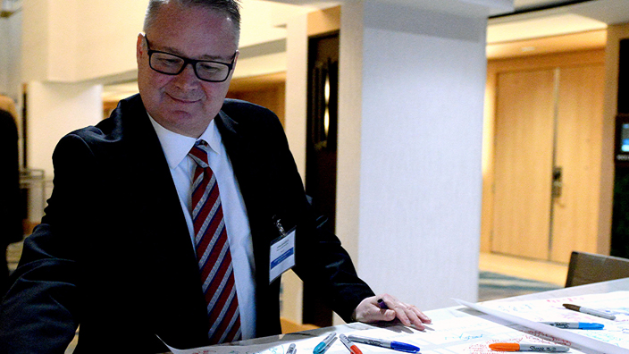 A man in a business suit and tie writes a message on a large piece of posterboard