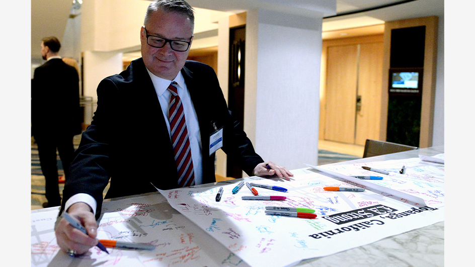 A man in a business suit and tie writes a message on a large piece of posterboard