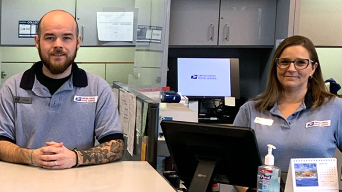 Three employees in USPS uniforms stand behind the retail counter in a Post Office