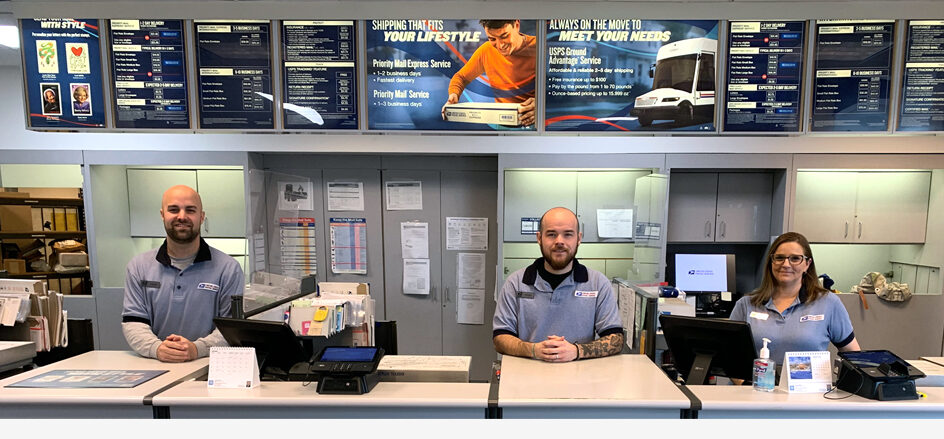 Three employees in USPS uniforms stand behind the retail counter in a Post Office