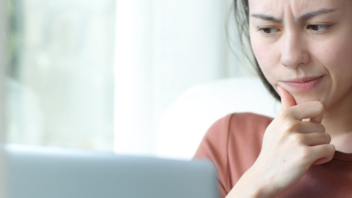 A woman sits in a living room using a laptop and casting a skeptical eye on the screen