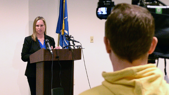 A woman stands at a lectern, addressing reporters and TV news crews