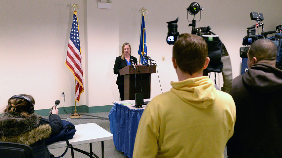 A woman stands at a lectern, addressing reporters and TV news crews