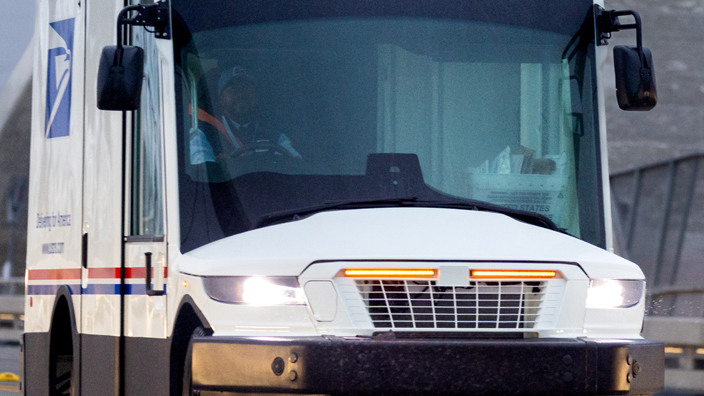 A modern USPS mail vehicle travels on a busy road