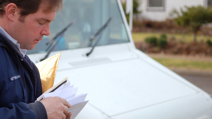 A man in a USPS uniform looks at a handful of mail while standing near a delivery vehicle