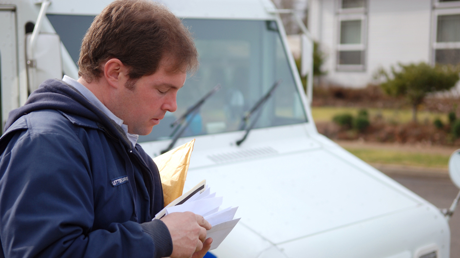 A man in a USPS uniform looks at a handful of mail while standing near a delivery vehicle