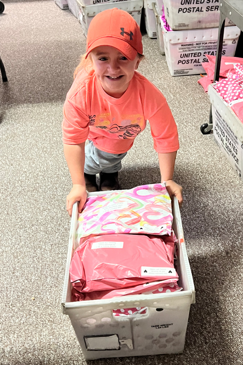 A young person holds a USPS crate full of colorful envelopes