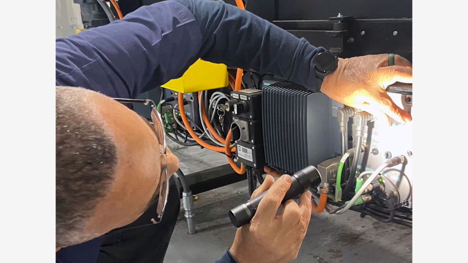 A man in a work shirt points a flashlight into mechanical equipment while holding a screw driver in another hand