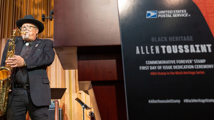 A man stands on stage and plays the saxophone in the background, near a podium in the foreground that bears Allen Toussaint stamp signage