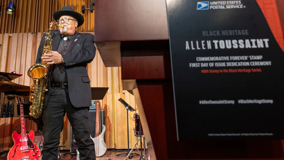 A man stands on stage and plays the saxophone in the background, near a podium in the foreground that bears Allen Toussaint stamp signage