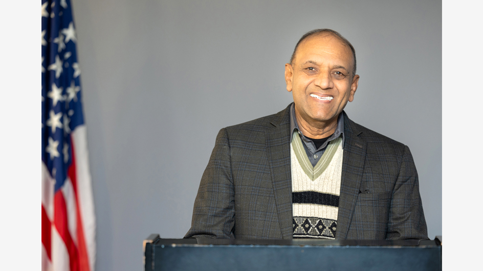 A man wearing a jacket and a sweater vest stands behind a lectern next to an American flag.