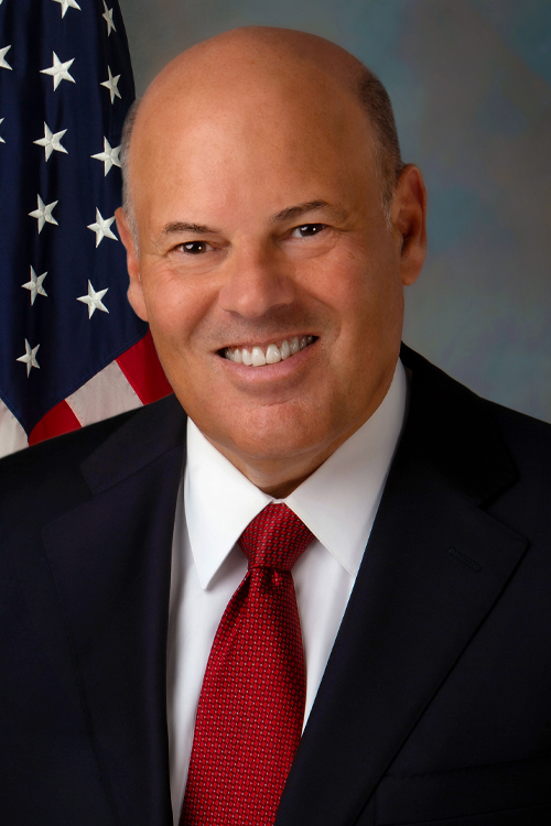 A studio portrait of a smiling man in a business suit, seated in front of a U.S. flag