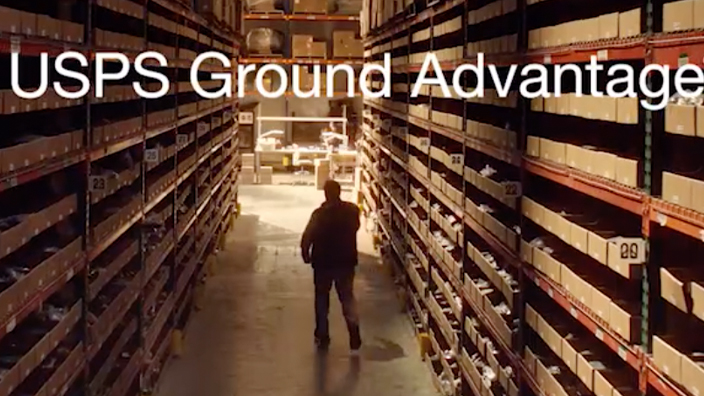 A wide shot of a warehouse stacked with boxes overshadowing worker, seen in silhouette. The USPS logo and the words “USPS Ground Advantage” appear in the center.