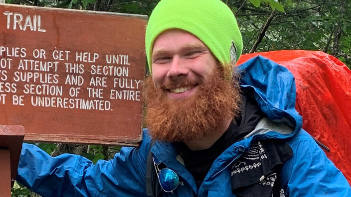 A young man wearing hiking gear stands near a brown marker bearing the words “Appalachian Trail”