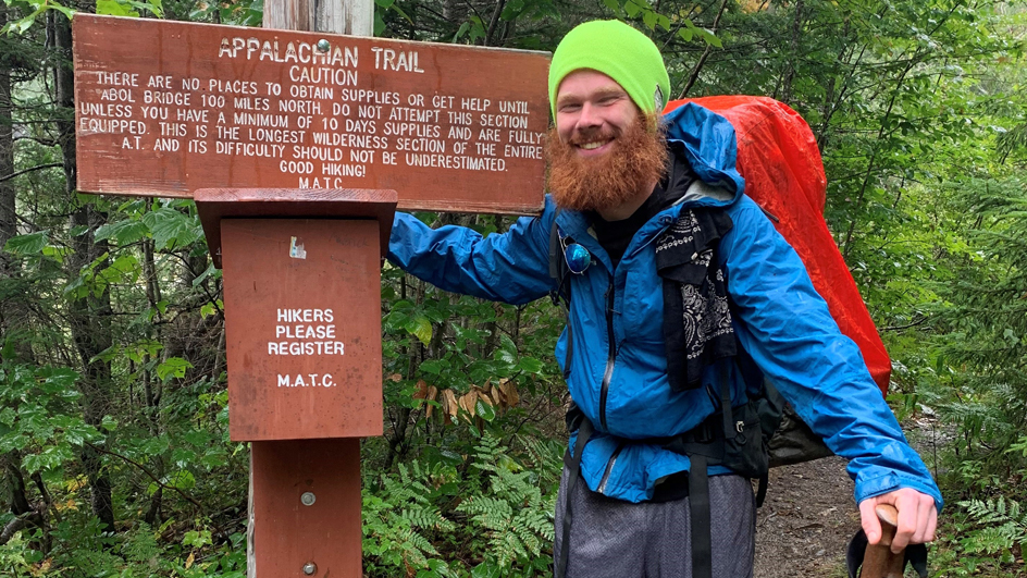 A young man wearing hiking gear stands near a brown marker bearing the words “Appalachian Trail”