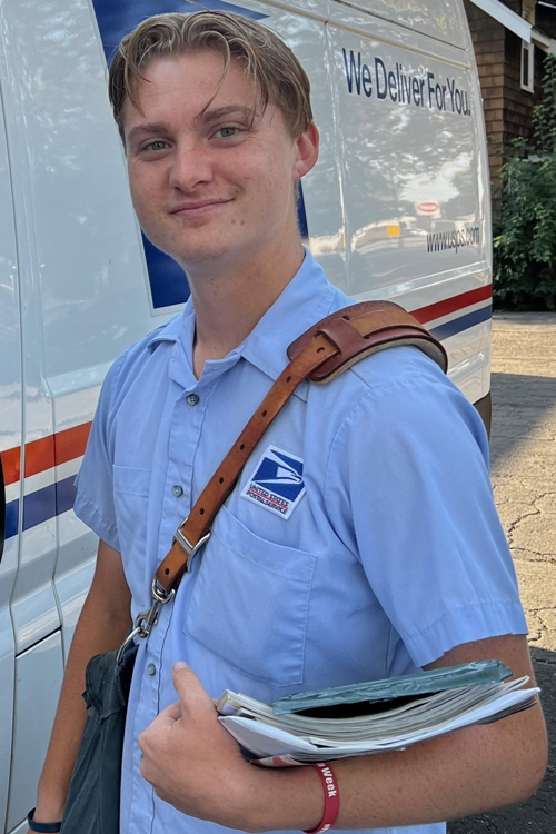 A man wearing a Postal Service uniform and a mail satchel over his shoulder stands next to a USPS delivery vehicle and smiles