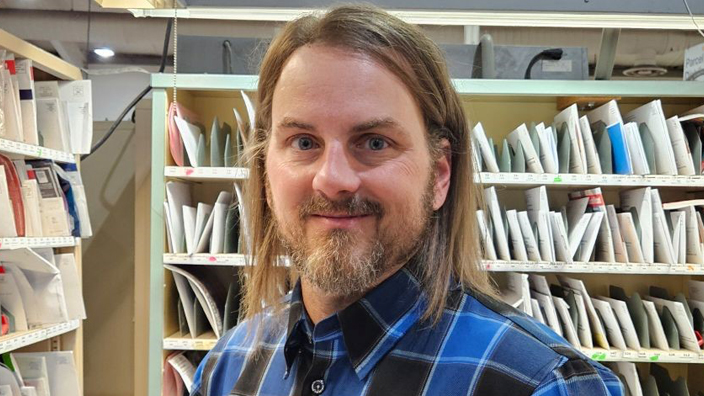 A man with a beard stands in front of a mail sorting rack