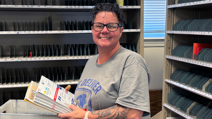 A woman wearing a postal-themed T-shirt smiles while standing in a Post Office workroom