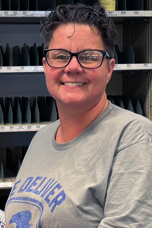 A woman wearing a postal-themed T-shirt smiles while standing in a Post Office workroom