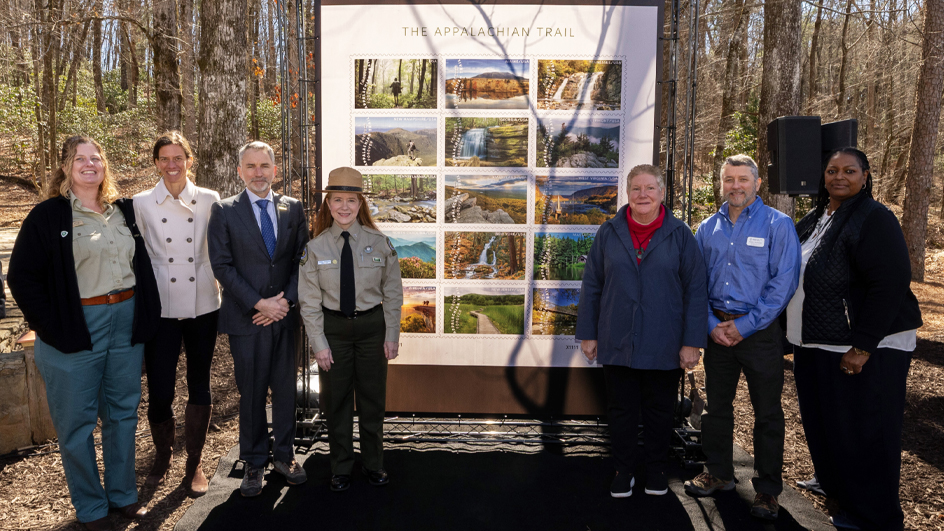 People stand in a forested setting, near a large poster displaying images of natural scenery.