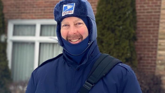 A man wearing heavy USPS-branded winter wear stands on a snow-covered residential street, holding a mail satchel