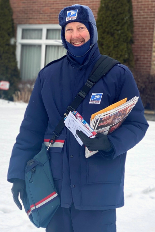 A man wearing heavy USPS-branded winter wear stands on a snow-covered residential street, holding a mail satchel