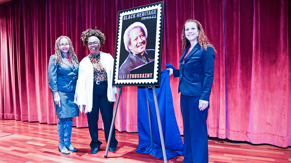 Three women in business wear stand on a stage next to a poster bearing an image of the Allen Toussaint stamp