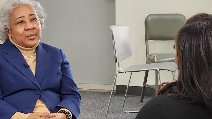 A Black woman dressed in a business suit sits in a chair and speaks to a TV reporter whose back is to the camera