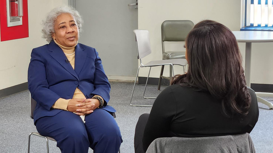 A Black woman dressed in a business suit sits in a chair and speaks to a TV reporter whose back is to the camera